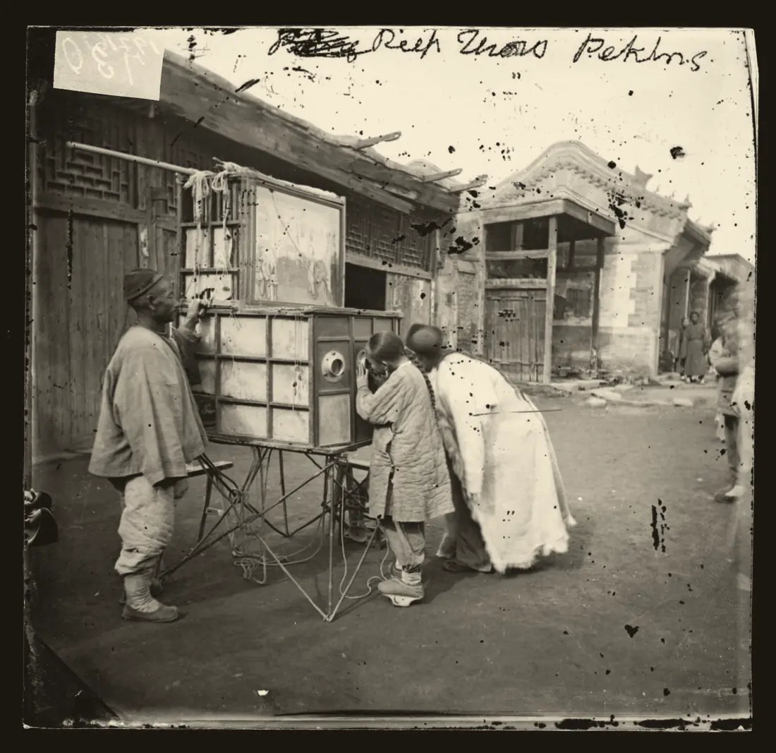 约翰·汤姆森 1869 年在北京拍的拉洋片:摊主手扶画箱站在一旁,一个梳辫子的孩子和一个披白袍的人弯腰凑在镜孔前,画箱顶上立着画片